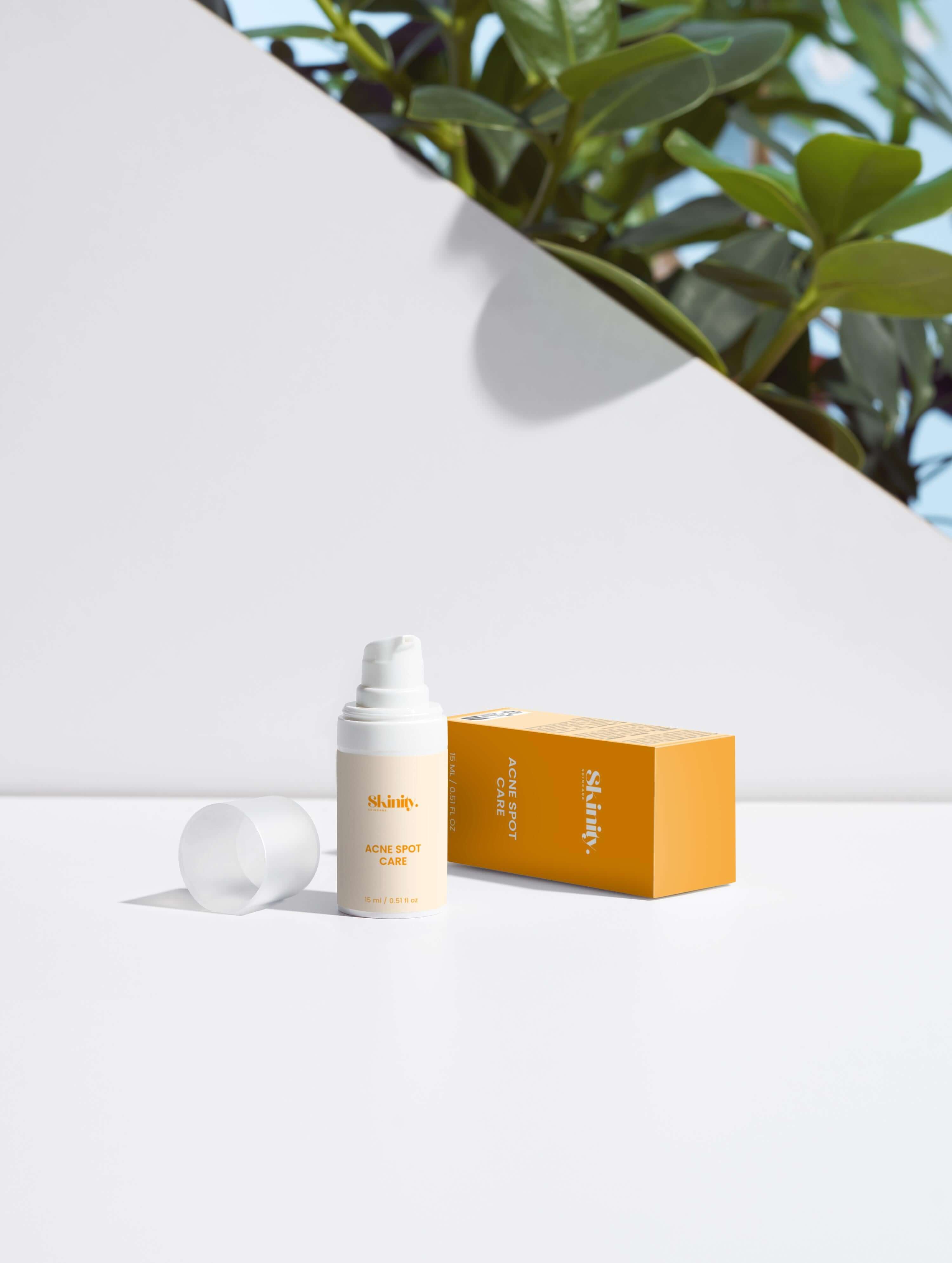 Acne spot care serum for all skin types next to its packaging on a white surface with greenery in the background.